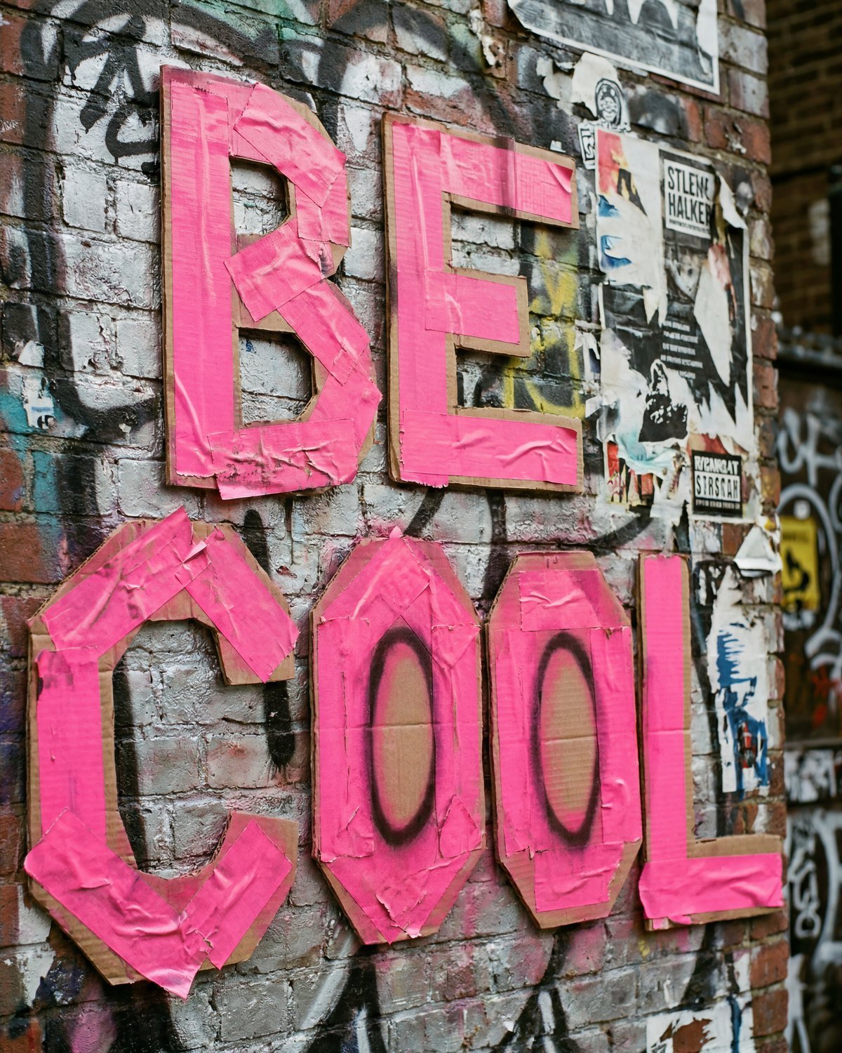 Hot-pink cardboard letters spelling BE COOL taped to a graffiti-covered brick wall